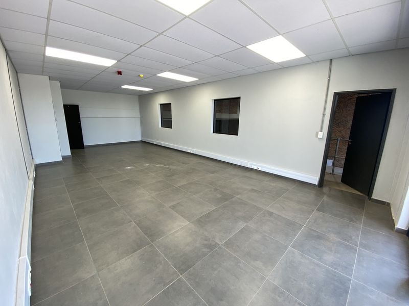 A spacious, empty industrial-style office featuring tiled flooring, a suspended ceiling with recessed lighting, and exposed brick accents near the entrance.