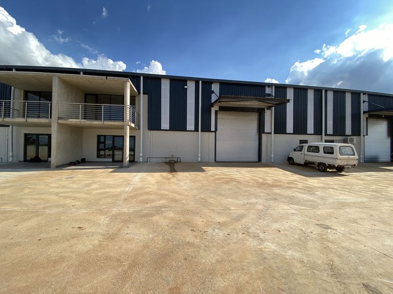 Industrial warehouse exterior featuring a large concrete yard, multiple roller shutter doors, and an integrated office balcony with steel railings.