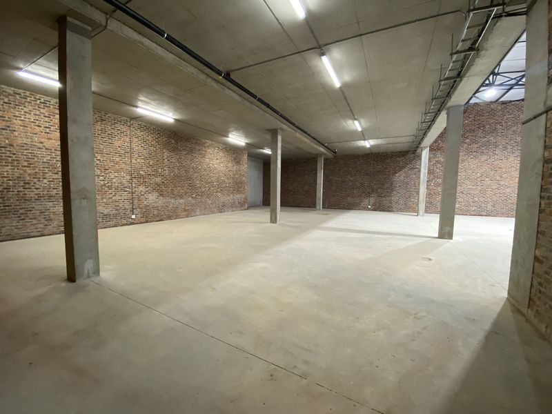 Spacious industrial warehouse area featuring exposed brick walls, concrete flooring, and high ceilings with functional strip lighting, suitable for various commercial or storage uses.