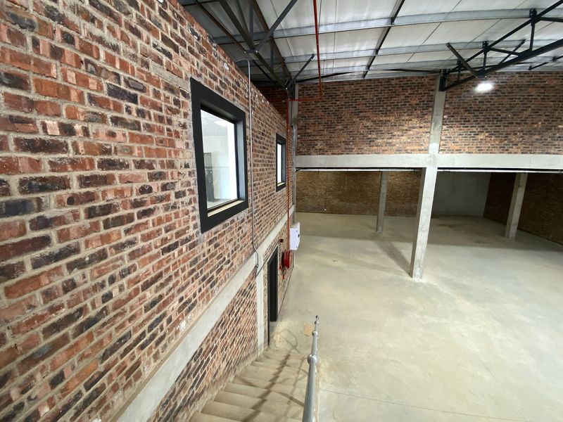 Spacious industrial warehouse interior featuring exposed brick walls, high ceilings with steel trusses, concrete flooring, and a mezzanine level overlooking the main floor.