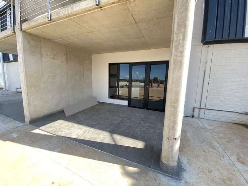A covered industrial-style patio featuring concrete structural elements, dark tiled flooring, and modern aluminium-framed glass doors leading into the building.