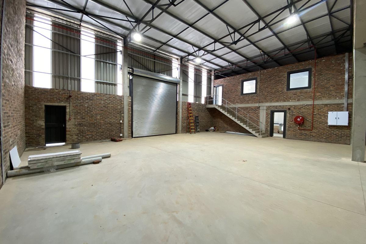 Spacious industrial warehouse unit featuring exposed brick walls, high ceilings with steel trusses, a large roller shutter door, and an internal staircase.