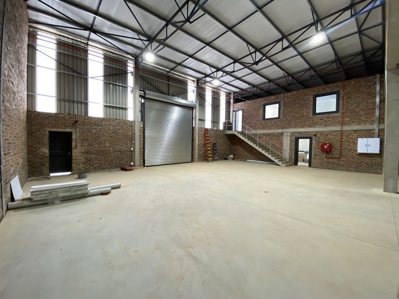 Spacious industrial warehouse unit featuring exposed brick walls, high ceilings with steel trusses, a large roller shutter door, and an internal staircase.