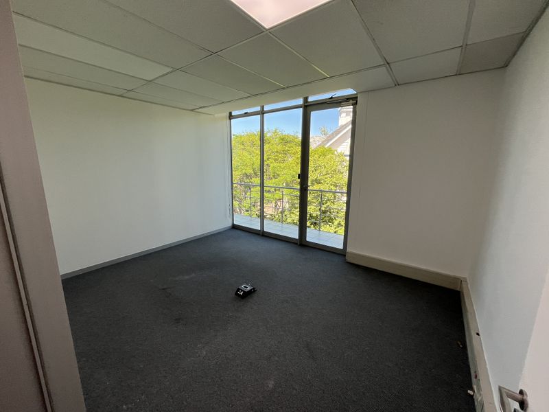 An empty office space featuring a suspended ceiling, carpeted flooring, and a glass door providing access to a small balcony with exterior views.