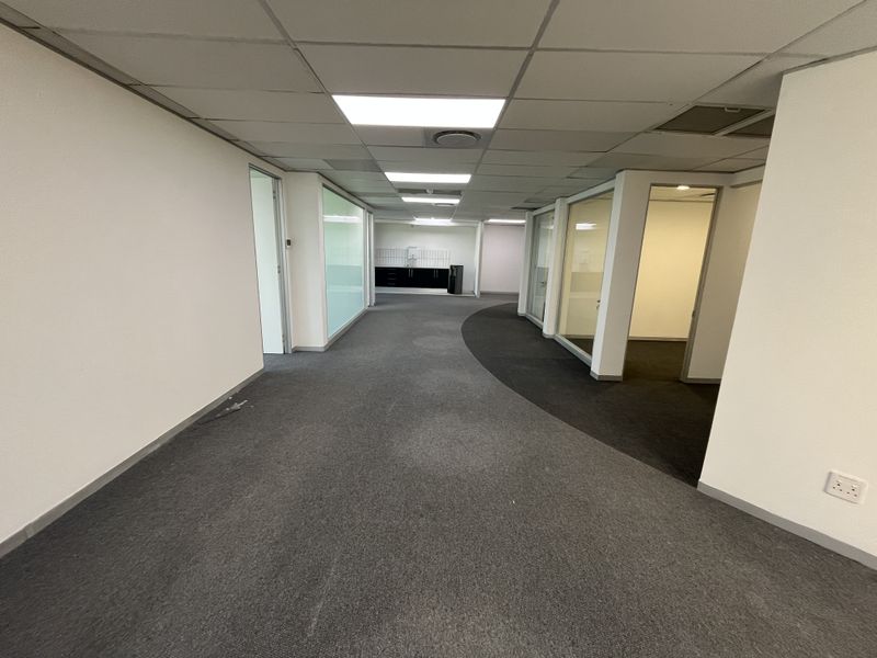Spacious office corridor featuring glass-partitioned rooms, suspended ceilings with recessed lighting, and a small built-in kitchenette area at the end of the hall.