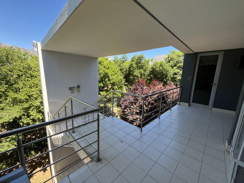 A spacious covered balcony featuring tiled flooring, a stainless steel balustrade, and direct access to an external staircase with surrounding greenery.