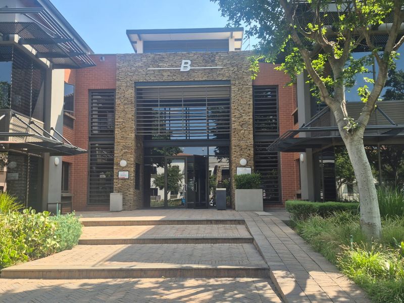 Front elevation of a modern office building featuring stone and brick facade, glass entrance, and paved approach with landscaped surroundings.