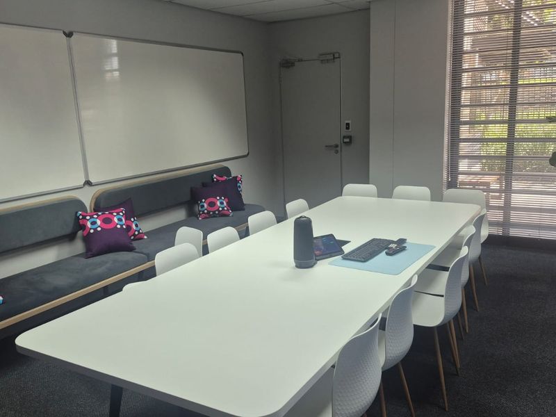 A modern office meeting room featuring built-in bench seating, wall-mounted whiteboards, and large windows with venetian blinds, providing a functional workspace.