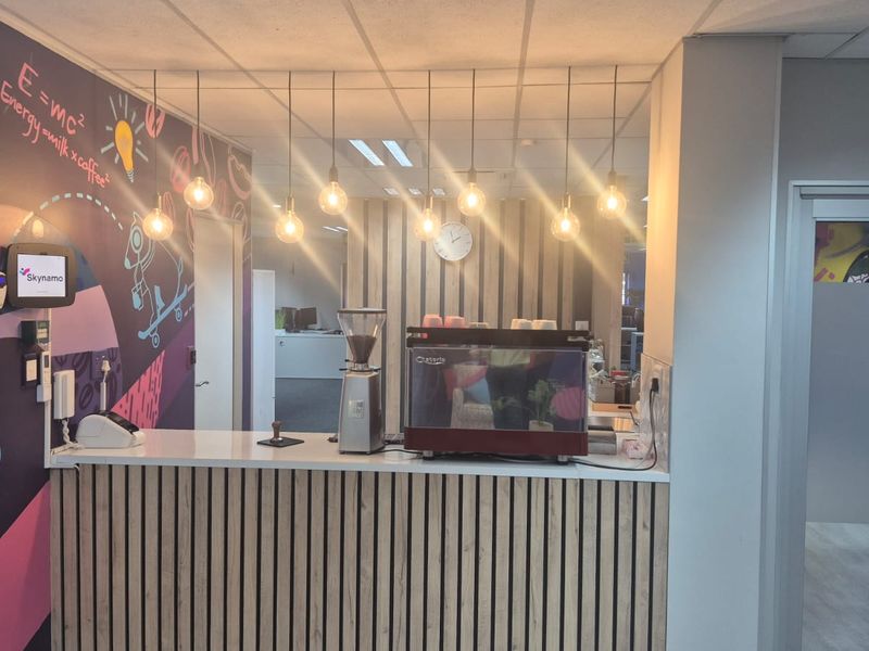 A modern office coffee station featuring a built-in counter with slatted wood detailing, pendant lighting, and a professional coffee machine setup.