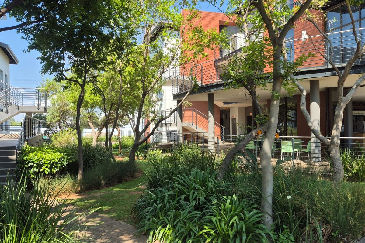 A well-maintained office complex exterior featuring a landscaped garden, paved walkways, and covered patio areas with modern brick and metal architectural elements.