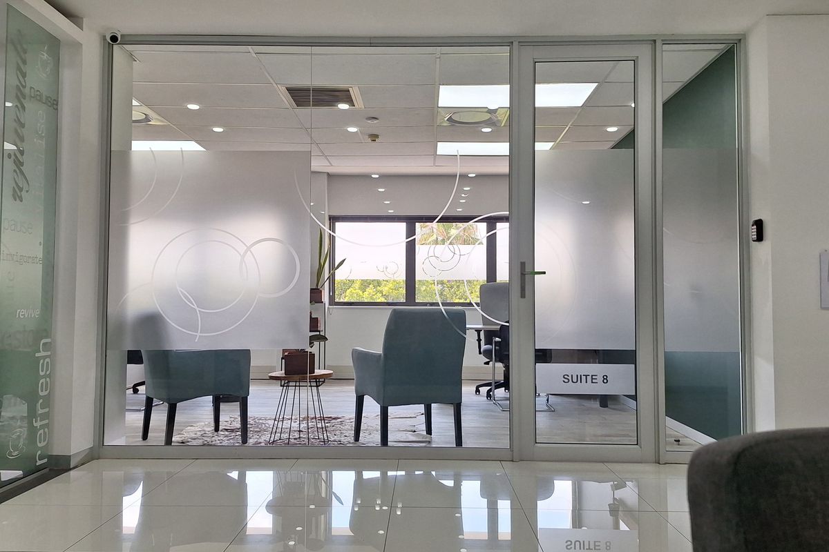 A modern office suite featuring a glass partition wall with frosted privacy film, tiled flooring, and recessed ceiling lighting.