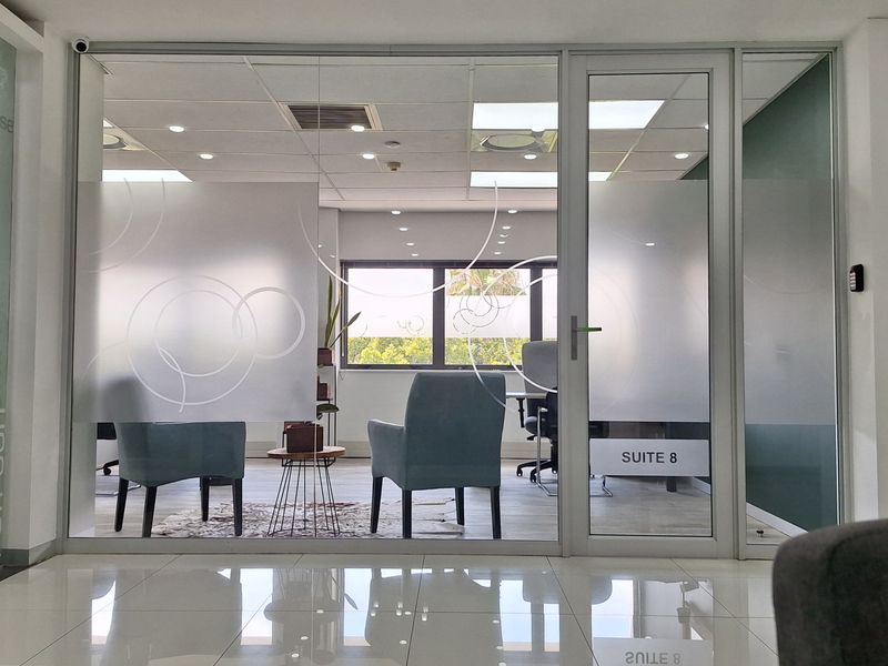 A modern office suite featuring a glass partition wall with frosted privacy film, tiled flooring, and recessed ceiling lighting.
