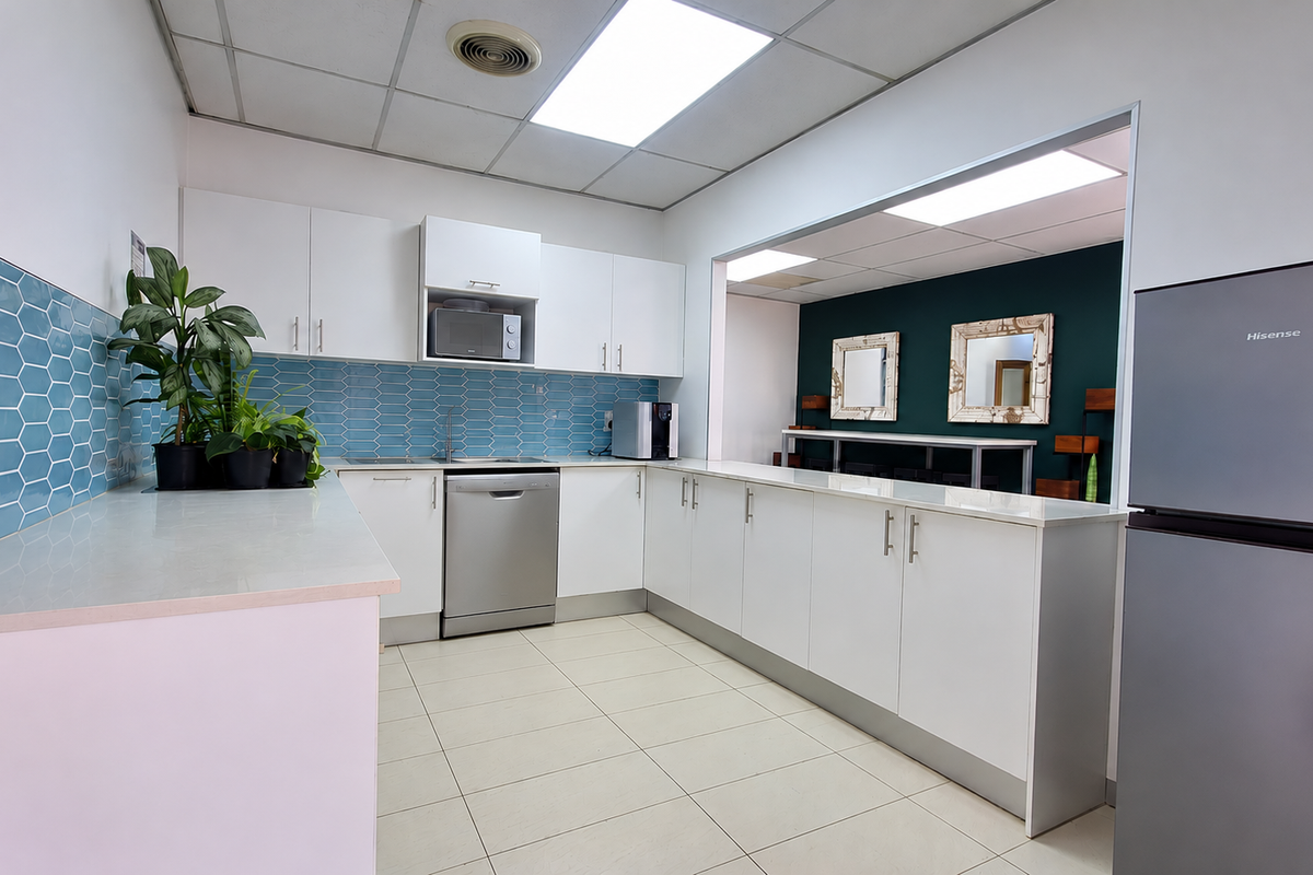 A functional office kitchen featuring white cabinetry, a blue hexagonal tiled backsplash, tiled flooring, and a pass-through window to an adjacent workspace.