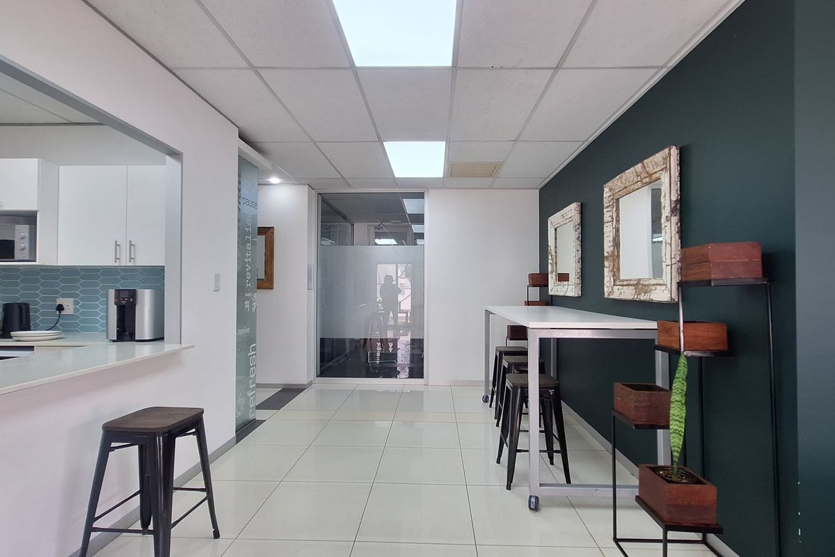 Modern open-plan office break area featuring a kitchenette with tiled backsplash, sleek cabinetry, and a bright, professional layout with glass partitions.