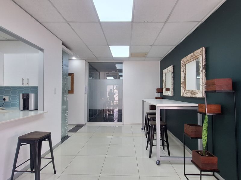Modern open-plan office break area featuring a kitchenette with tiled backsplash, sleek cabinetry, and a bright, professional layout with glass partitions.