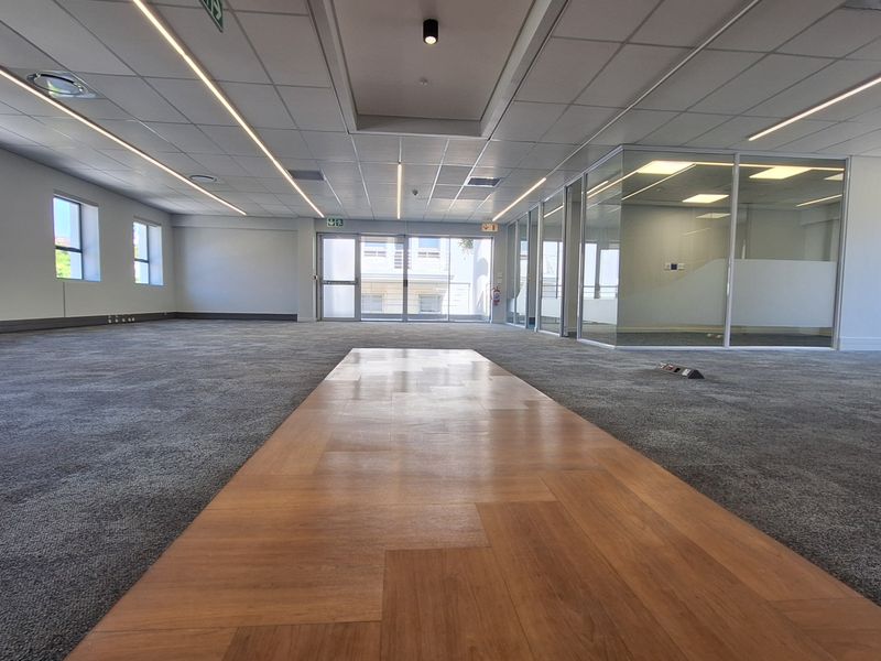 A spacious, modern open-plan office area featuring a mix of carpet and wood laminate flooring, glass partitioned offices, and contemporary recessed linear lighting.