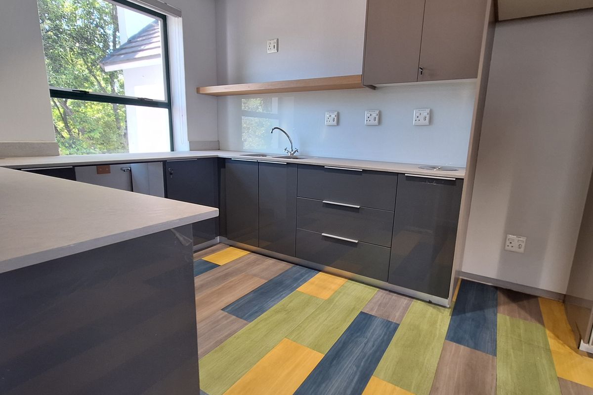 A modern, empty office kitchenette featuring sleek grey cabinetry, stone countertops, a stainless steel sink, and a vibrant multi-colored vinyl floor.
