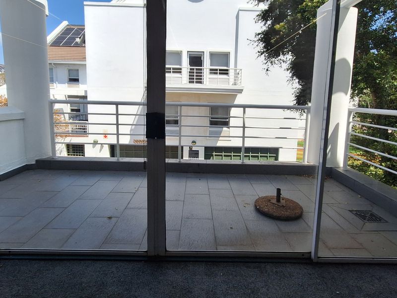 A clean, tiled balcony featuring a white metal balustrade and sliding glass door access, overlooking the surrounding office complex.
