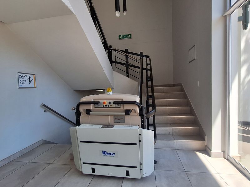 Bright office entrance area featuring a tiled staircase with a built-in wheelchair platform lift and large glass windows providing natural light.