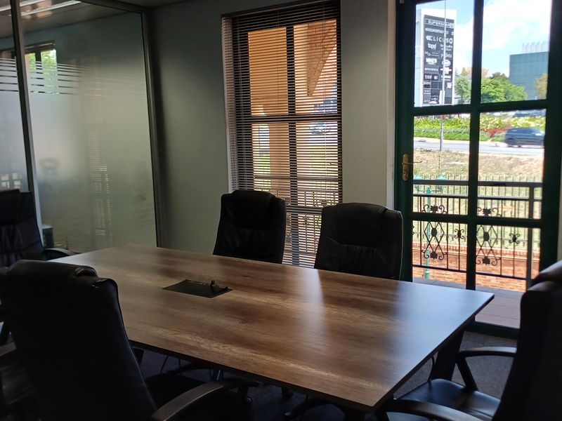 A professional office meeting room featuring a glass partition wall, venetian blinds, and a framed glass door with exterior views.