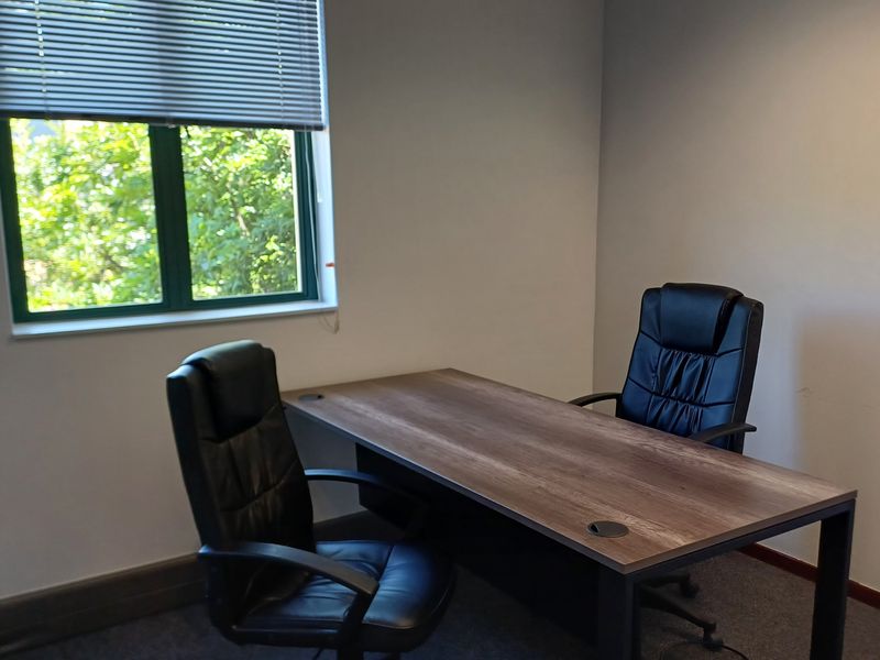 A functional office space featuring carpeted flooring, neutral walls, and a window with venetian blinds providing natural light.