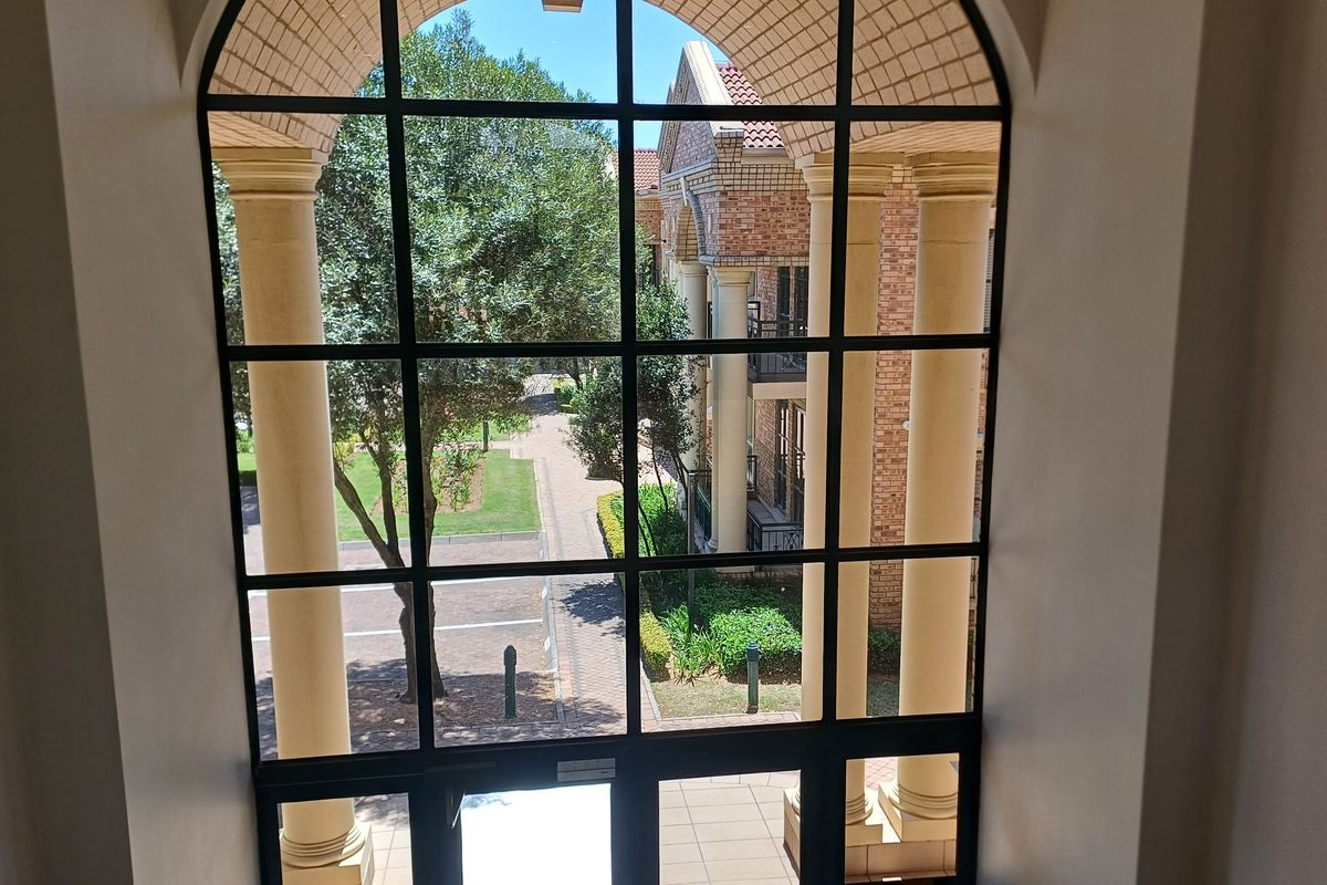 A view through a large arched window featuring black metal framing and classical columns, overlooking the landscaped office complex grounds.