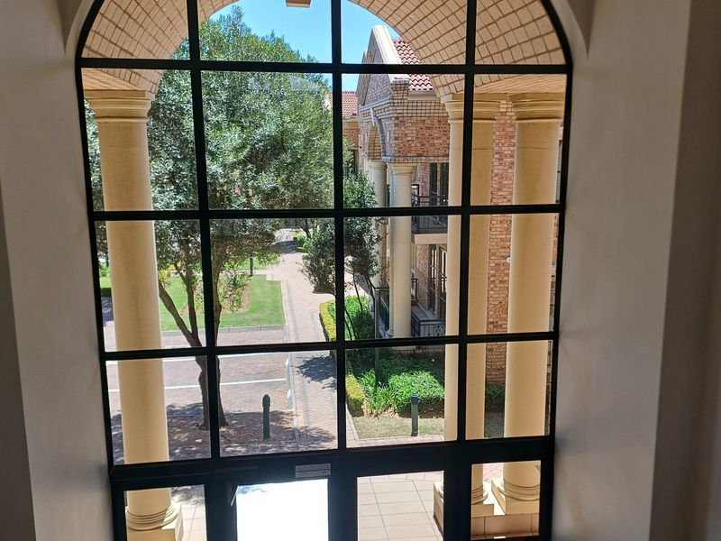 A view through a large arched window featuring black metal framing and classical columns, overlooking the landscaped office complex grounds.