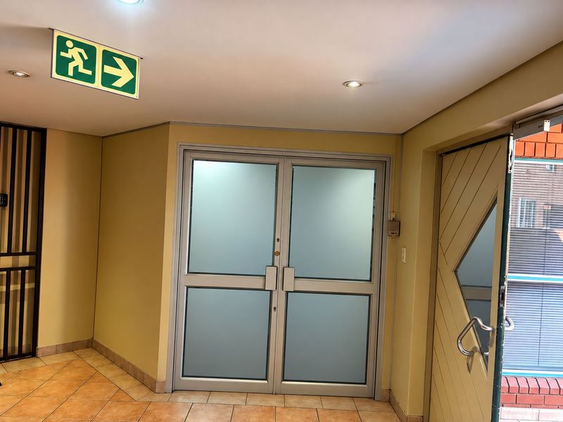 A clean and functional office entry area featuring frosted glass double doors, tiled flooring, and integrated emergency exit signage.