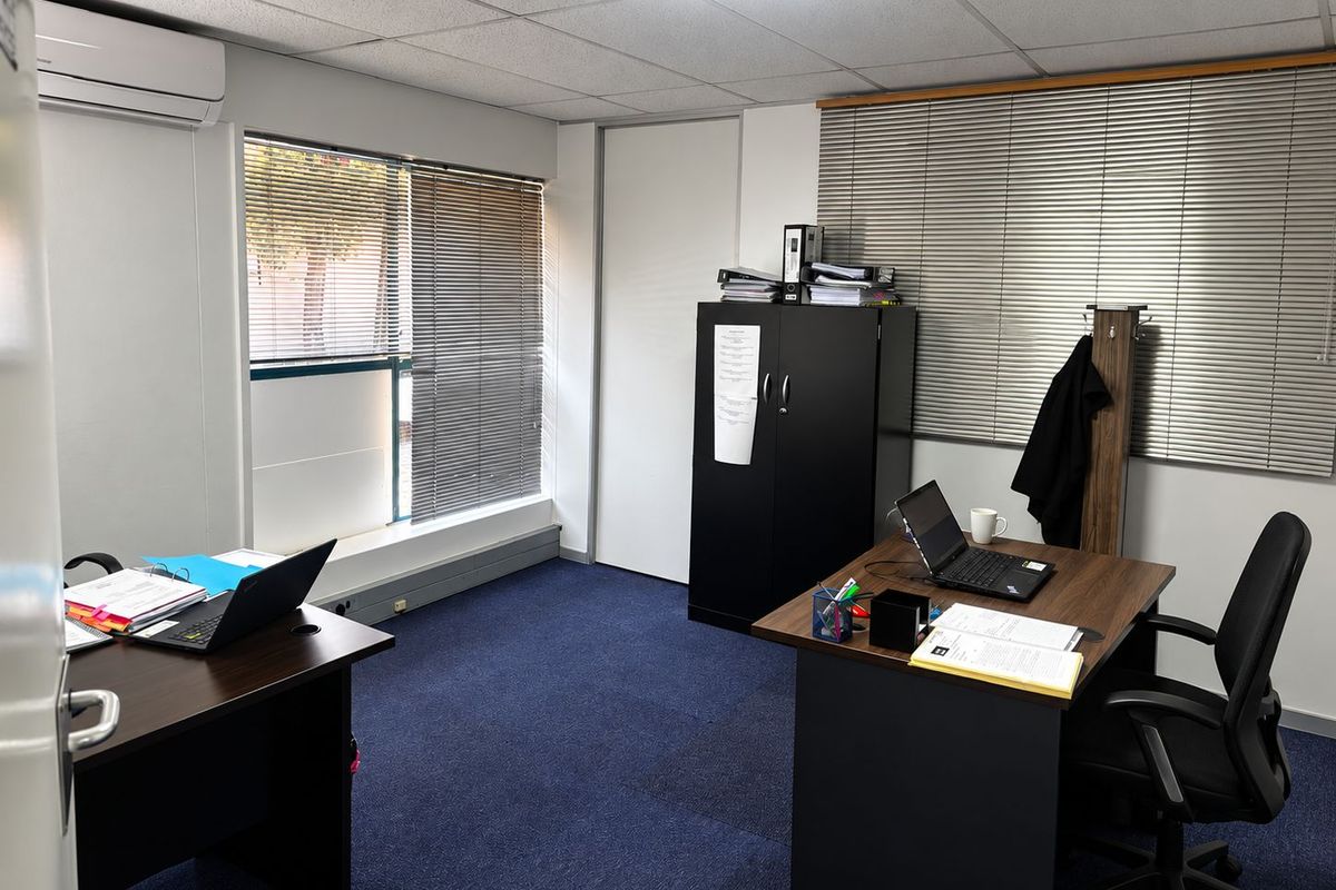 A functional office space featuring carpeted flooring, suspended ceiling tiles, wall-mounted air conditioning, and large windows with fitted venetian blinds.
