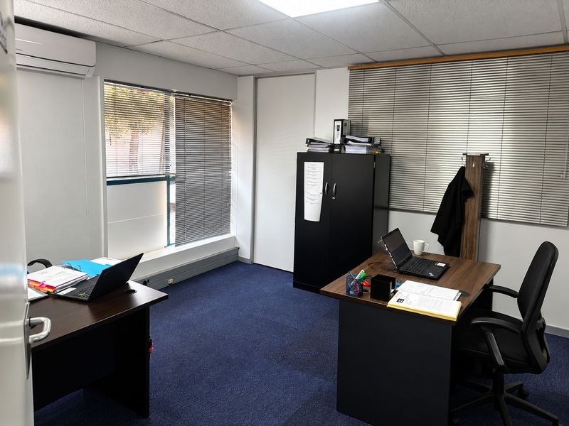 A functional office space featuring carpeted flooring, suspended ceiling tiles, wall-mounted air conditioning, and large windows with fitted venetian blinds.