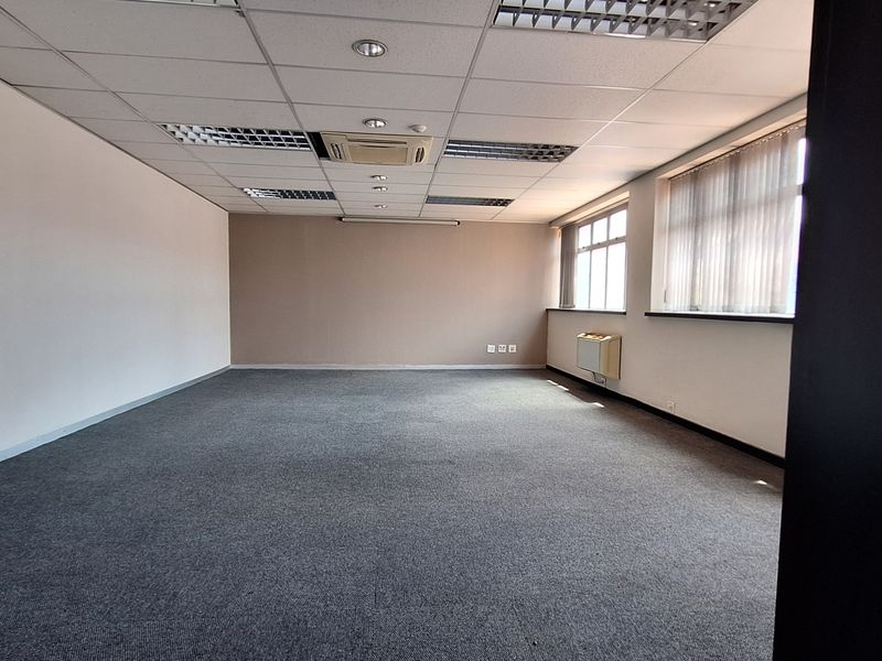 A spacious, empty office area featuring a suspended ceiling with recessed lighting, carpeted flooring, and dual air conditioning units.
