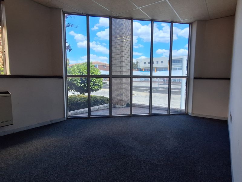 An empty office space featuring large floor-to-ceiling windows, carpeted flooring, and a wall-mounted HVAC unit, offering a bright and functional workspace.