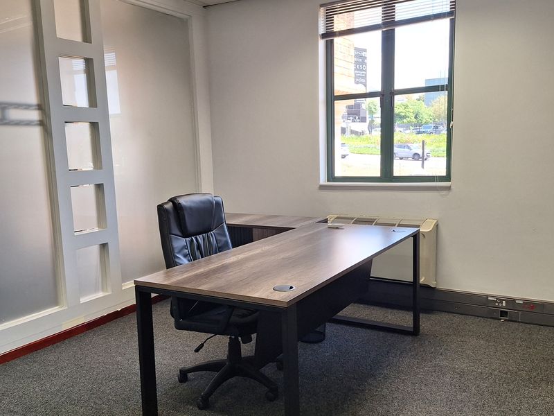 A professional office space featuring carpeted flooring, a frosted glass partition, and a window with blinds, providing a functional work environment.