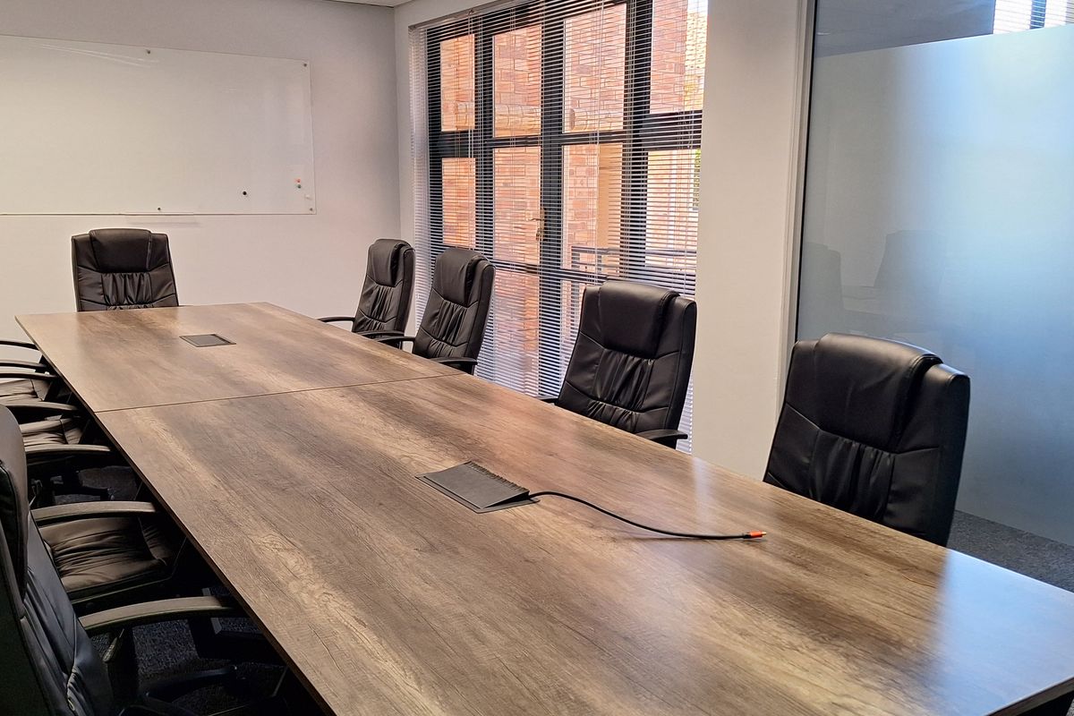 A professional office boardroom featuring a large meeting table, glass partition walls, suspended ceiling with recessed lighting, and large windows with blinds.