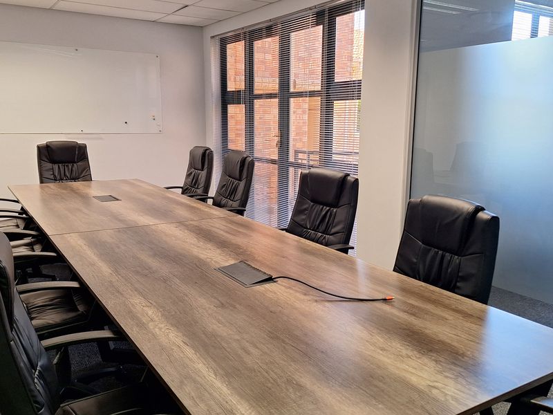 A professional office boardroom featuring a large meeting table, glass partition walls, suspended ceiling with recessed lighting, and large windows with blinds.