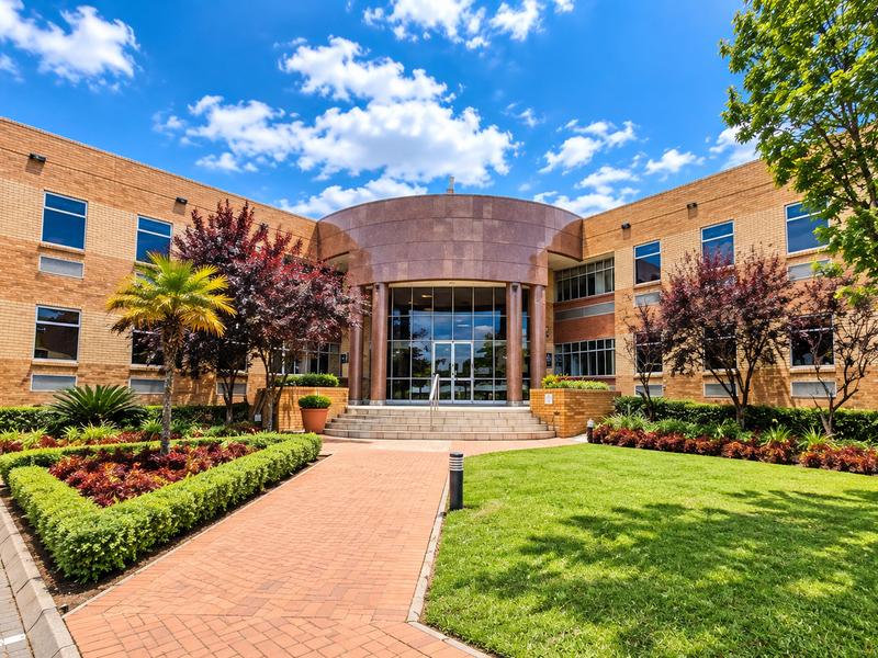 A professional exterior view of a modern brick office building featuring a central stone-clad entrance, glass facade, and well-maintained landscaped gardens.