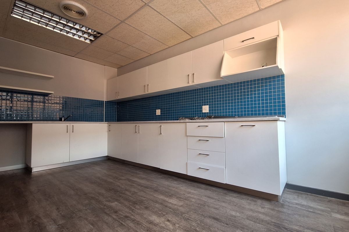 A clean, empty office kitchenette featuring white built-in cabinetry, blue tiled backsplash, laminate flooring, and a drop ceiling with recessed lighting.