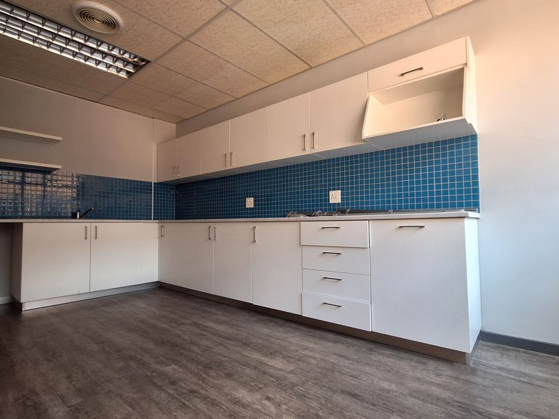 A clean, empty office kitchenette featuring white built-in cabinetry, blue tiled backsplash, laminate flooring, and a drop ceiling with recessed lighting.