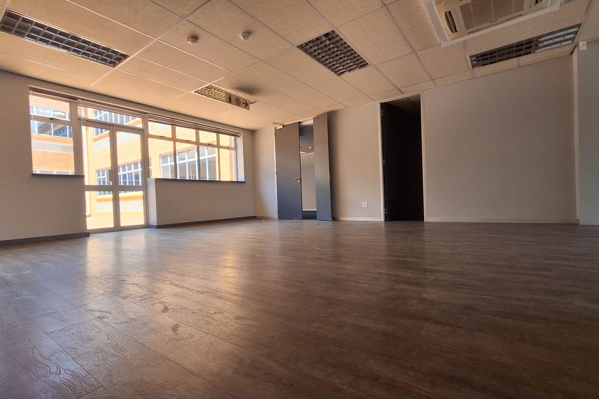 A spacious, empty office area featuring laminate flooring, large windows for natural light, and a suspended ceiling with integrated lighting and air conditioning.