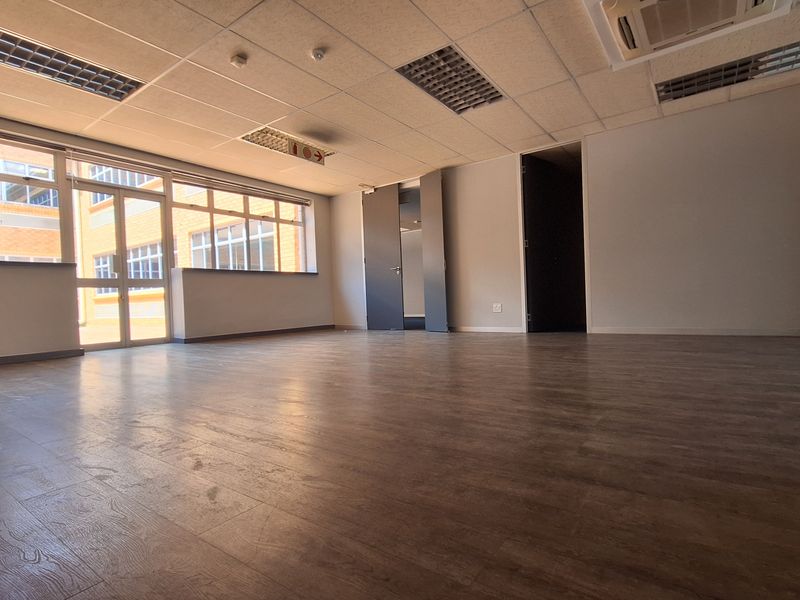 A spacious, empty office area featuring laminate flooring, large windows for natural light, and a suspended ceiling with integrated lighting and air conditioning.