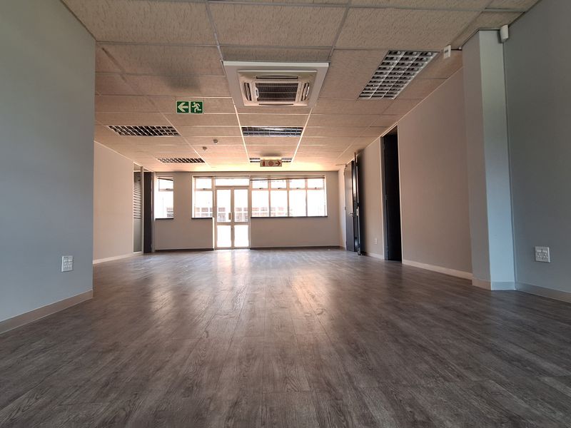A spacious, vacant office area featuring modern laminate flooring, a suspended ceiling with integrated lighting, and a cassette air conditioning unit.