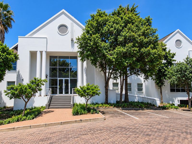 A professional exterior view of a modern office building featuring a white facade, large glass entrance, paved driveway, and well-maintained landscaped surroundings.