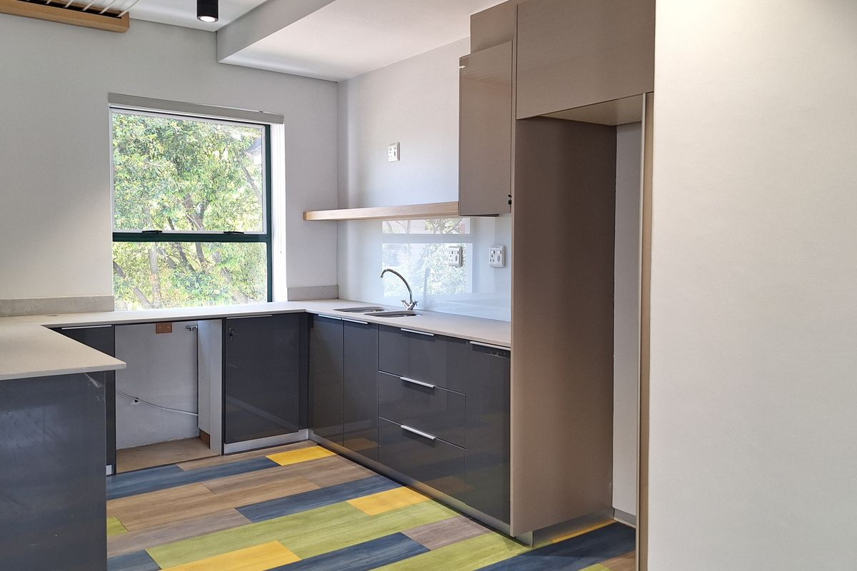 A modern, empty office kitchen featuring sleek grey cabinetry, light countertops, a glass splashback, and a stainless steel sink with a mixer tap.