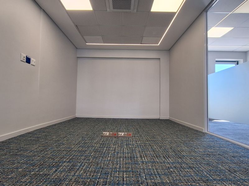 A modern, empty office space featuring carpeted flooring, a suspended ceiling with recessed lighting, and a glass partition wall.
