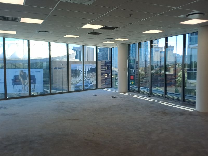 Spacious, empty open-plan office area featuring floor-to-ceiling windows with city views, suspended ceilings, and carpeted flooring.
