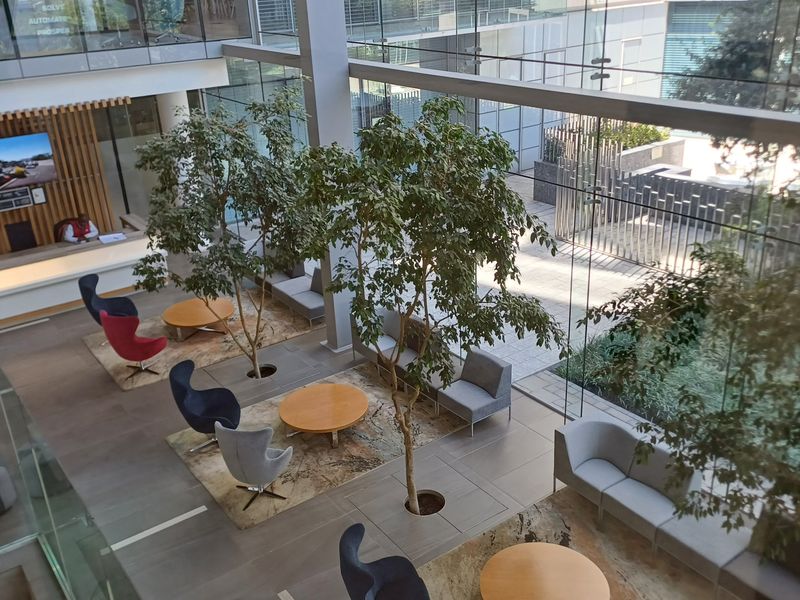 Modern office building atrium featuring floor-to-ceiling glass walls, a reception desk, and integrated indoor landscaping with trees.