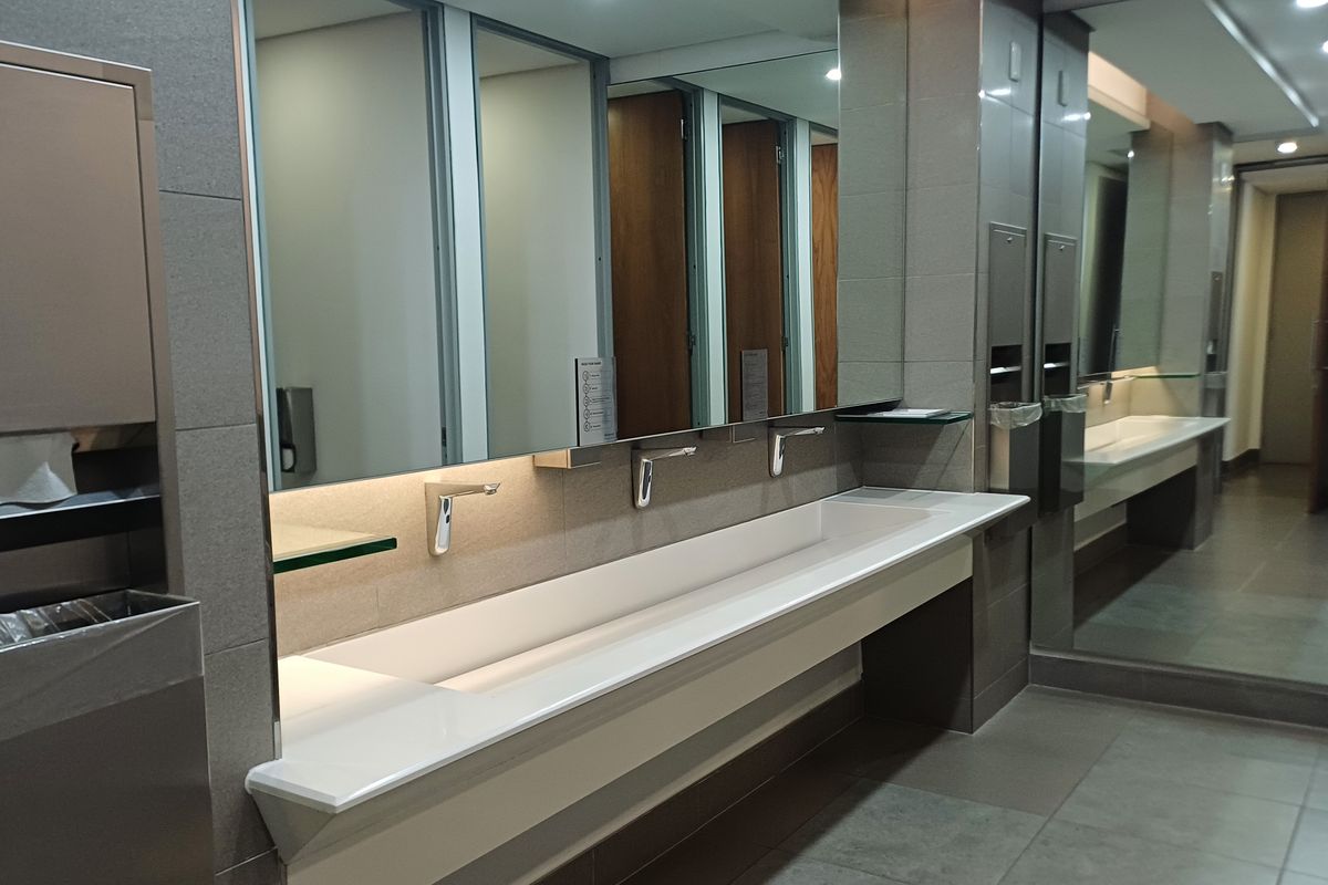 Modern commercial office bathroom featuring a sleek integrated trough sink, wall-mounted sensor taps, and large vanity mirrors with recessed lighting.