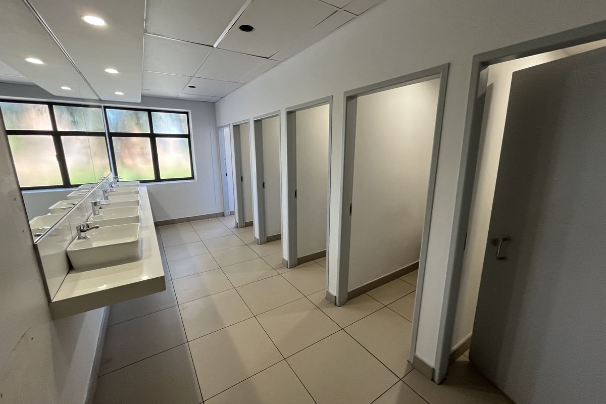 A functional office bathroom featuring multiple vanity basins, tiled flooring, and several private toilet cubicles with a suspended ceiling.