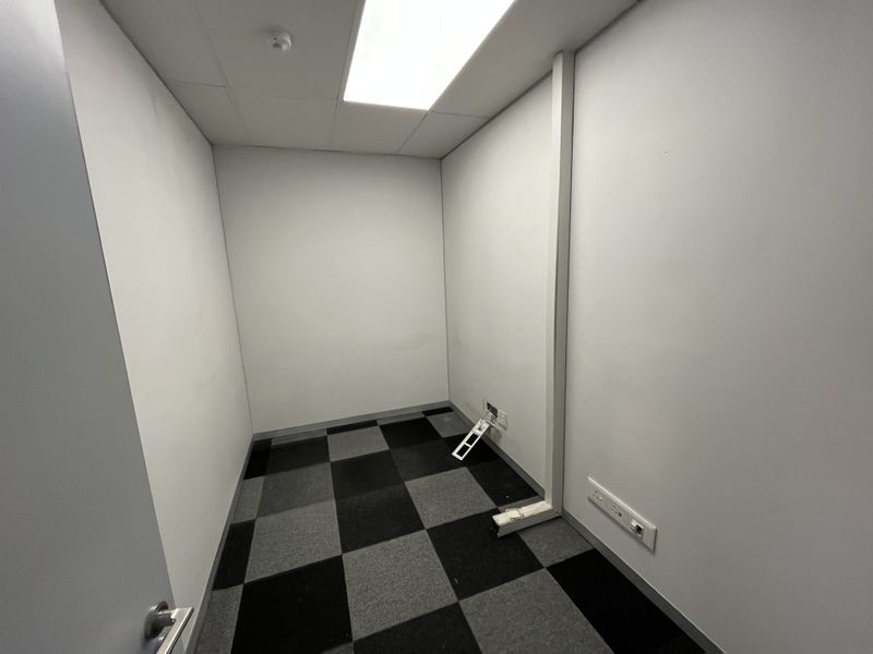 A small, empty office space featuring a suspended ceiling, recessed lighting, and checkered carpet tiles, with visible electrical trunking and power points.