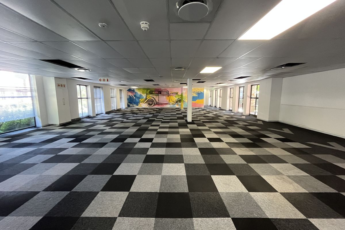 A large, open-plan office space featuring a checkered carpet tile floor, suspended ceiling with recessed lighting, and large windows providing natural light.